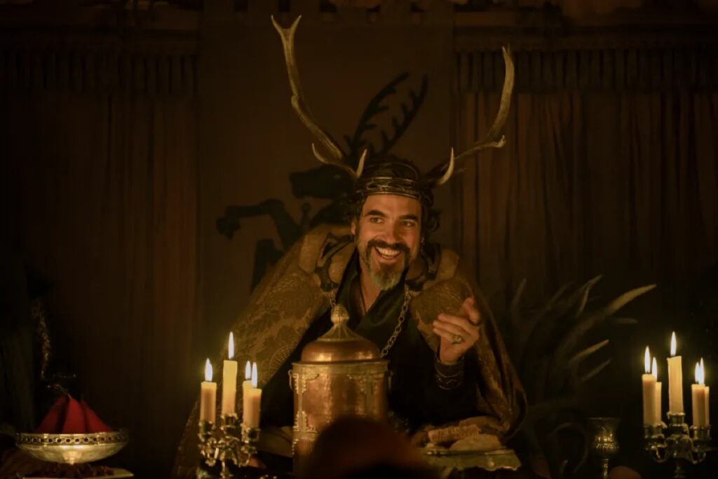 man with a dark beard at a feast, smiling and wearing an antler headdress.