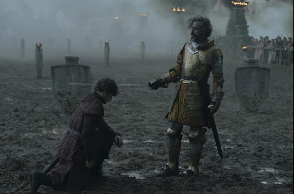 young man kneeling on a muddy jousting field as a standing knight taps a sword to his shoulder.