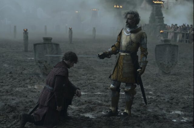 Một chàng trai trẻ đang quỳ trên một bãi đấu thương lầy lội, trong khi một hiệp sĩ đứng thẳng gõ kiếm vào vai anh ta.