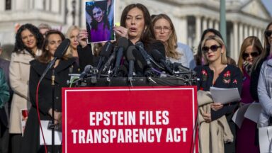 A woman speaking at a podium during a news conference holds up a picture of herself from when she was young. The podium has numerous microphones and a sign that says, "Epstein Files Transparency Act."