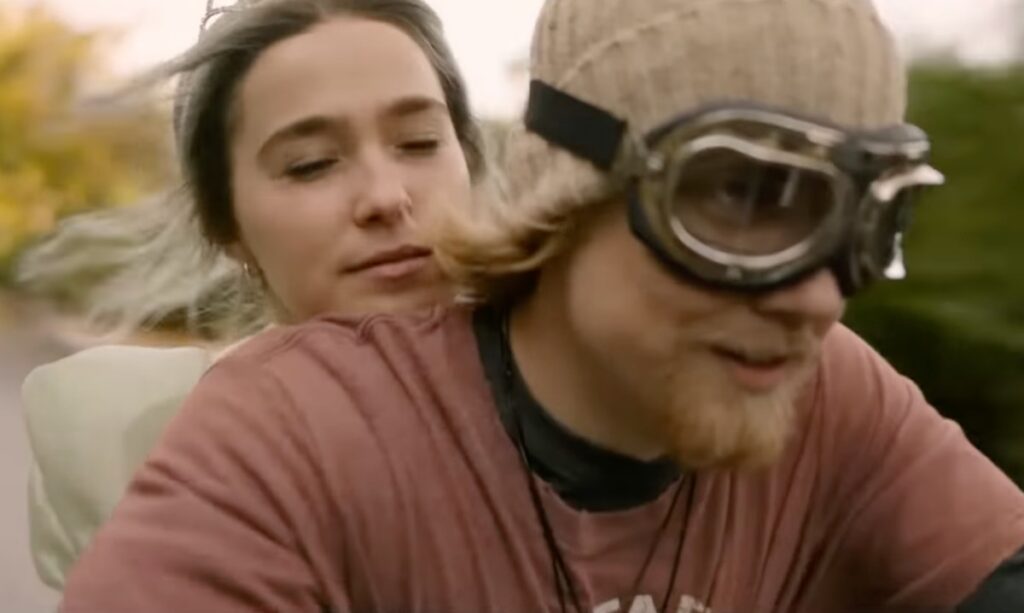 blonde woman clinging to back of man driving a motorcycle, looking blissful