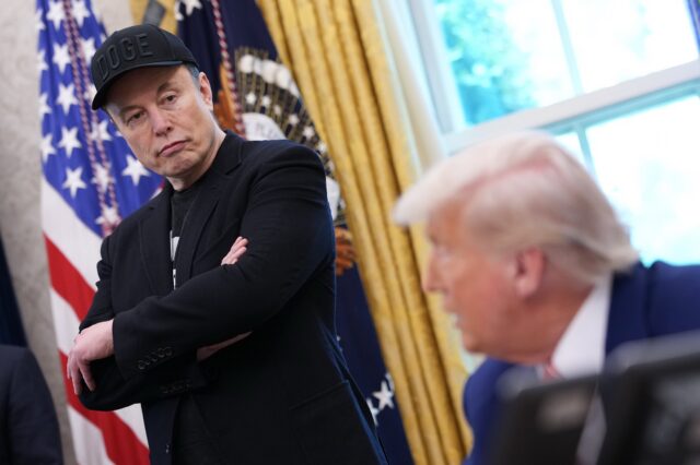 Elon Musk stands with his arms crossed, wearing a DOGE cap, in the Oval Office, near President Trump who sits at his desk while speaking.