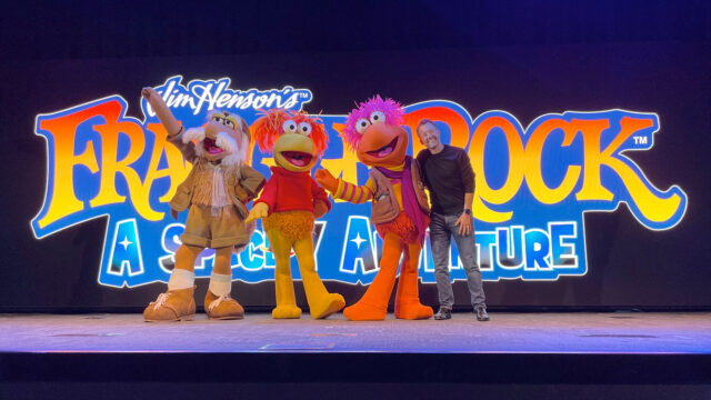 Costumed "walk-around" characters pose with a man in a dark shirt and jeans in front of a digitally displayed logo