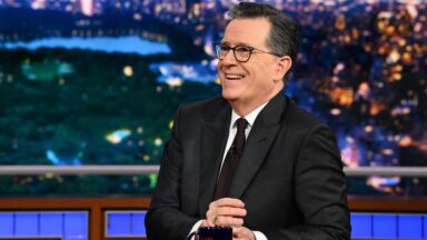 Stephen Colbert sits at his desk and smiles on the set of his late-night TV show.