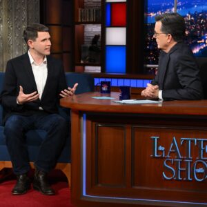 Stephen Colbert interviews a guest while sitting at his desk on the set of his late-night show.