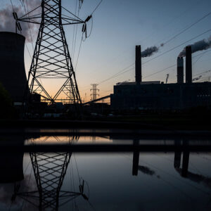 A bluish early dawn sky in the backdrop, with a dark set of buildings of a power plant, cooling towards, and transmission lines.