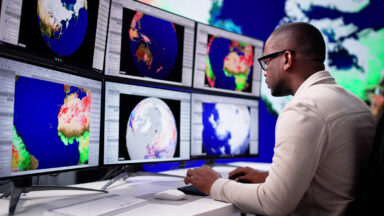 A man sits at a bank of computer screens, each showing different views of the planet.