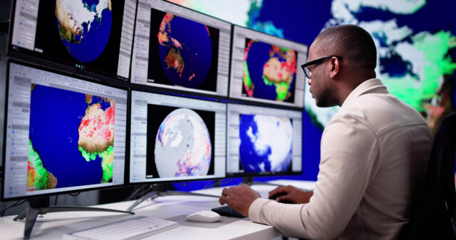 A man sits at a bank of computer screens, each showing different views of the planet.