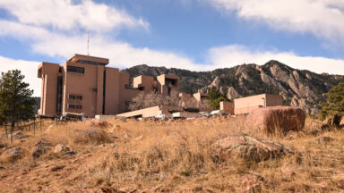 The National Center for Atmospheric Research in Boulder, Colorado on December 17, 2025. Clay colored building with grass and red rocks in foreground.