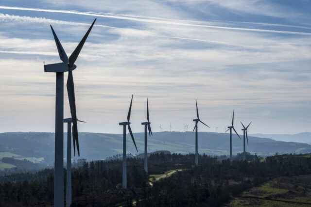Dry, shrub colored hillsides with a large series of wind turbines poking up into a sky filled with wispy clouds.