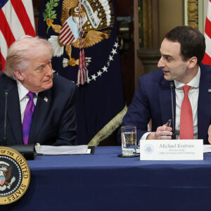 Two men turn to each other while seated behind a table, with several flags in the background.