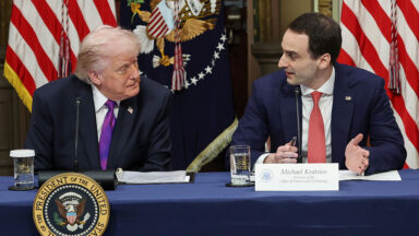 Two men turn to each other while seated behind a table, with several flags in the background.