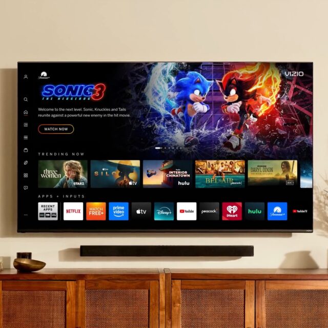 A Vizio smart TV mounted to a wall above a soundbar and wooden sideboard.