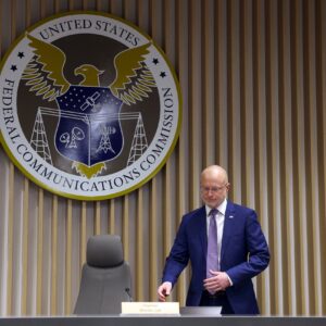 FCC Chairman Brendan Carr prepares to sit down for a meeting. A large FCC crest is seen on the wall above him.