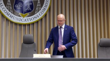 FCC Chairman Brendan Carr prepares to sit down for a meeting. A large FCC crest is seen on the wall above him.