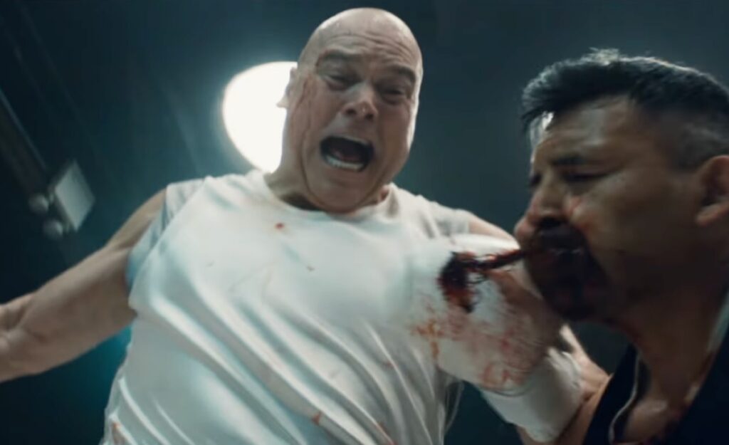 bald man in bloodied white tee-shirt angrily whaling on another moment in a fight ring. Blood is spurting out of the other man's mouth