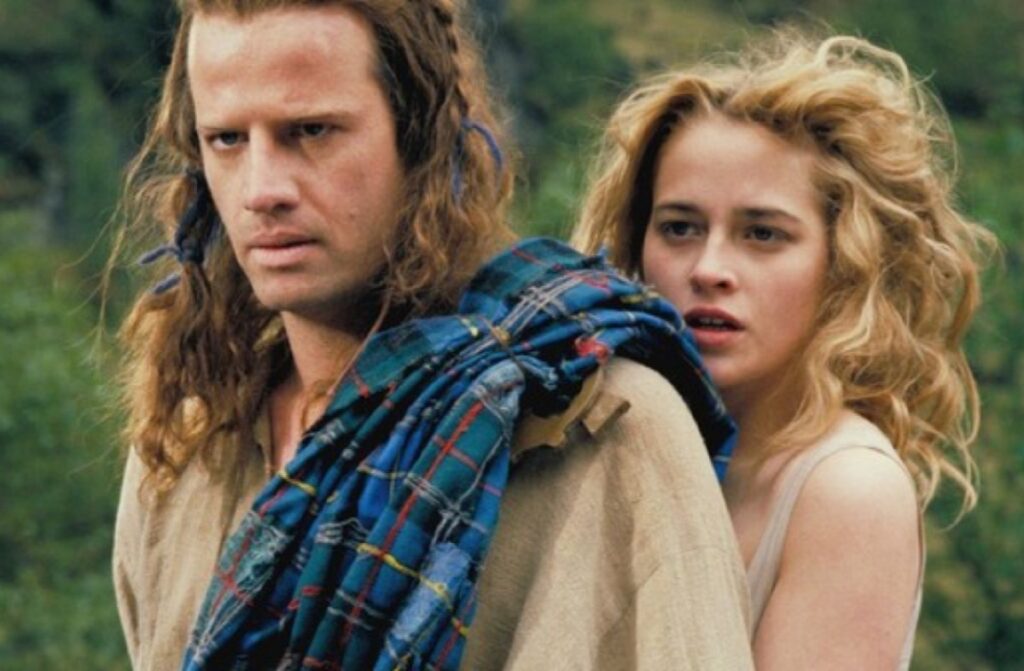 long haired scotsman in kilt stands protectively in front of pretty blonde woman.