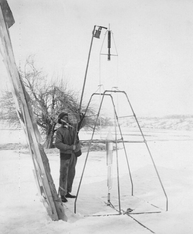 A black and white photo of a man holding a pole next to a early rocket on a snowy field.
