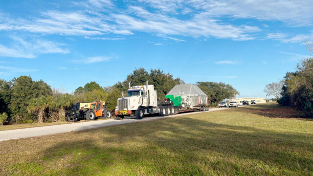 a truck tows an oversize payload down a one-lane road under a partially cloudy blue sky