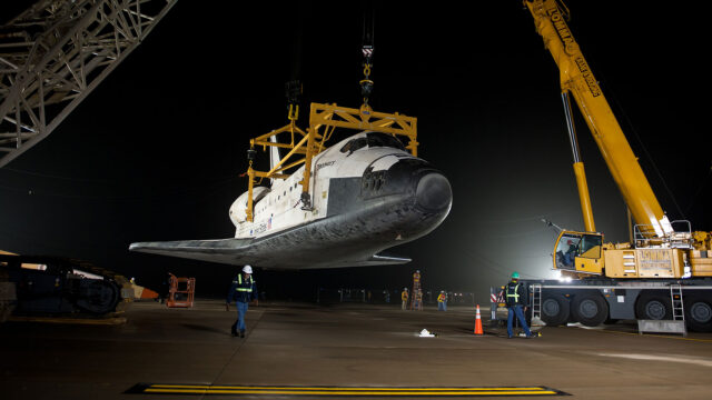 a black and white winged orbiter is hoisted by a bright yellow crane off the ground in the darkness of night, lit by a spotlight
