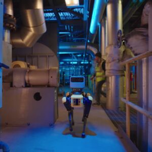 A four-legged Boston Dynamics robot called Spot walks down the hallway of an industrial facility. A human worker wearing a bright yellow safety vest is starting to cross the hallway behind the robot.