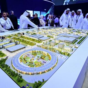 People wearing head coverings and long white robes are standing around a model of the largest data center in the UAE under construction in Abu Dhabi as the Stargate initiative, a joint venture between G42, Microsoft, and OpenAI, during the Abu Dhabi International Petroleum Exhibition Conference (ADIPEC) in Abu Dhabi on November 3, 2025.