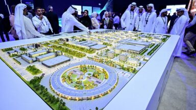 People wearing head coverings and long white robes are standing around a model of the largest data center in the UAE under construction in Abu Dhabi as the Stargate initiative, a joint venture between G42, Microsoft, and OpenAI, during the Abu Dhabi International Petroleum Exhibition Conference (ADIPEC) in Abu Dhabi on November 3, 2025.