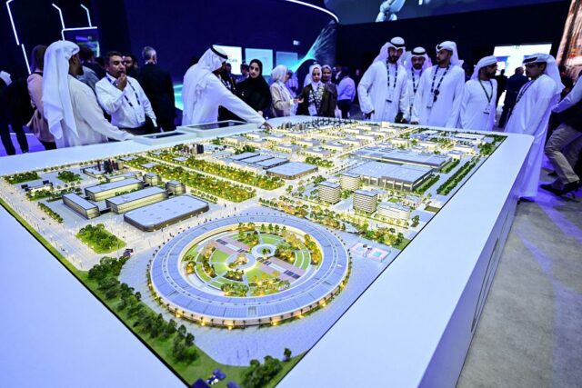 People wearing head coverings and long white robes are standing around a model of the largest data center in the UAE under construction in Abu Dhabi as the Stargate initiative, a joint venture between G42, Microsoft, and OpenAI, during the Abu Dhabi International Petroleum Exhibition Conference (ADIPEC) in Abu Dhabi on November 3, 2025.