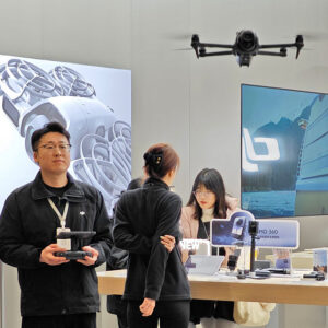 A DJI store employee wearing black clothing holds a drone controller as he pilots a quadcopter drone hovering in the foreground. Behind the man is a DJI store interior display with several other employees or customers in the background.