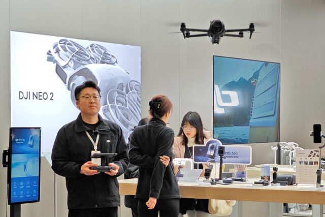 A DJI store employee wearing black clothing holds a drone controller as he pilots a quadcopter drone hovering in the foreground. Behind the man is a DJI store interior display with several other employees or customers in the background.