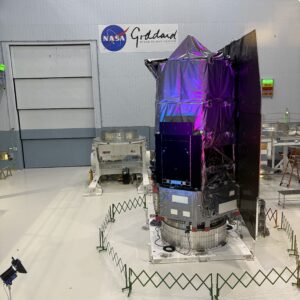 An enormous room with a NASA-Goddard banner on one wall. In the center sits a structure with a small fence around it. The structure is wrapped in dark fabric, and roughly cylindrical. It is large enough to extend above the second floor of the building.