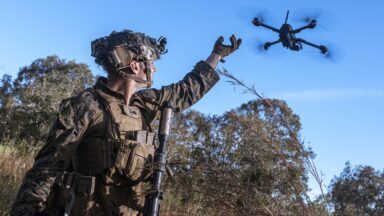 Wearing a camouflaged uniform and helmet, Marine Corps Cpl. Calvin Burke holds his arm outstretched as he activates a small drone to survey the defensive line for opposing forces during a simulated assault and seizure at Glen Airfield, Queensland, Australia, July 2025.