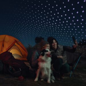 People camping at night take a selfie with a dog, using a phone connected to a satellite network.