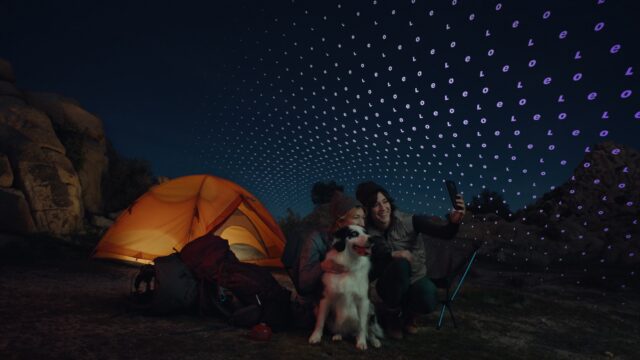 People camping at night take a selfie with a dog, using a phone connected to a satellite network.