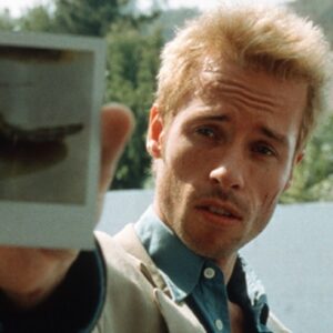 Man with cropped blond hair and a haggard look holding up a developed Polaroid