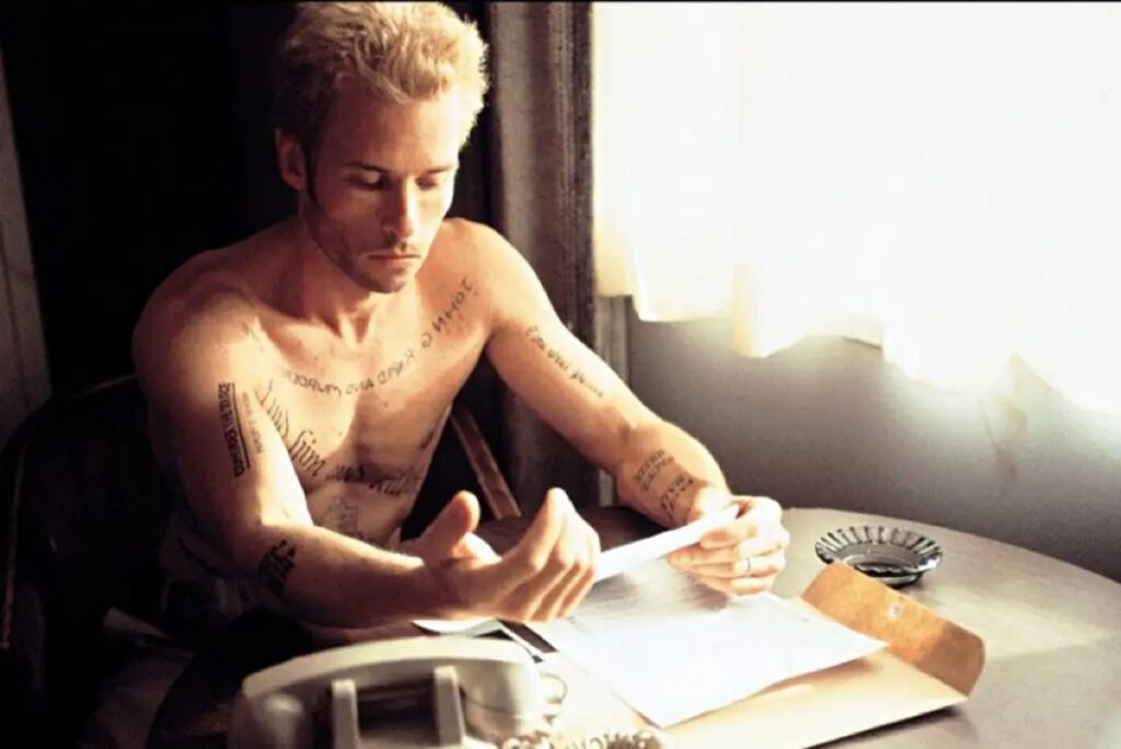 shirtless man with cropped bleached blond hair sitting at a table poring over notes