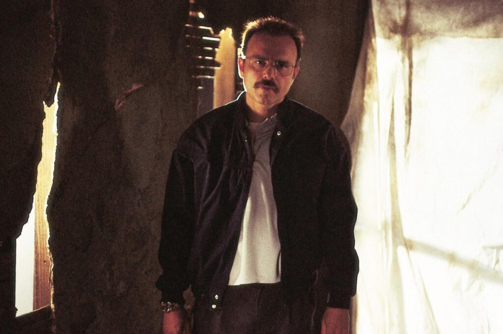 Man with a mustache and glasses standing in a decrepit room
