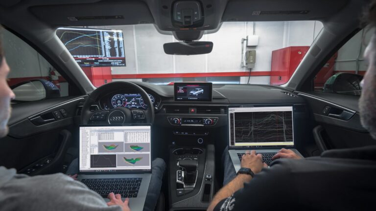Two men with laptops sit in the front seats of a car