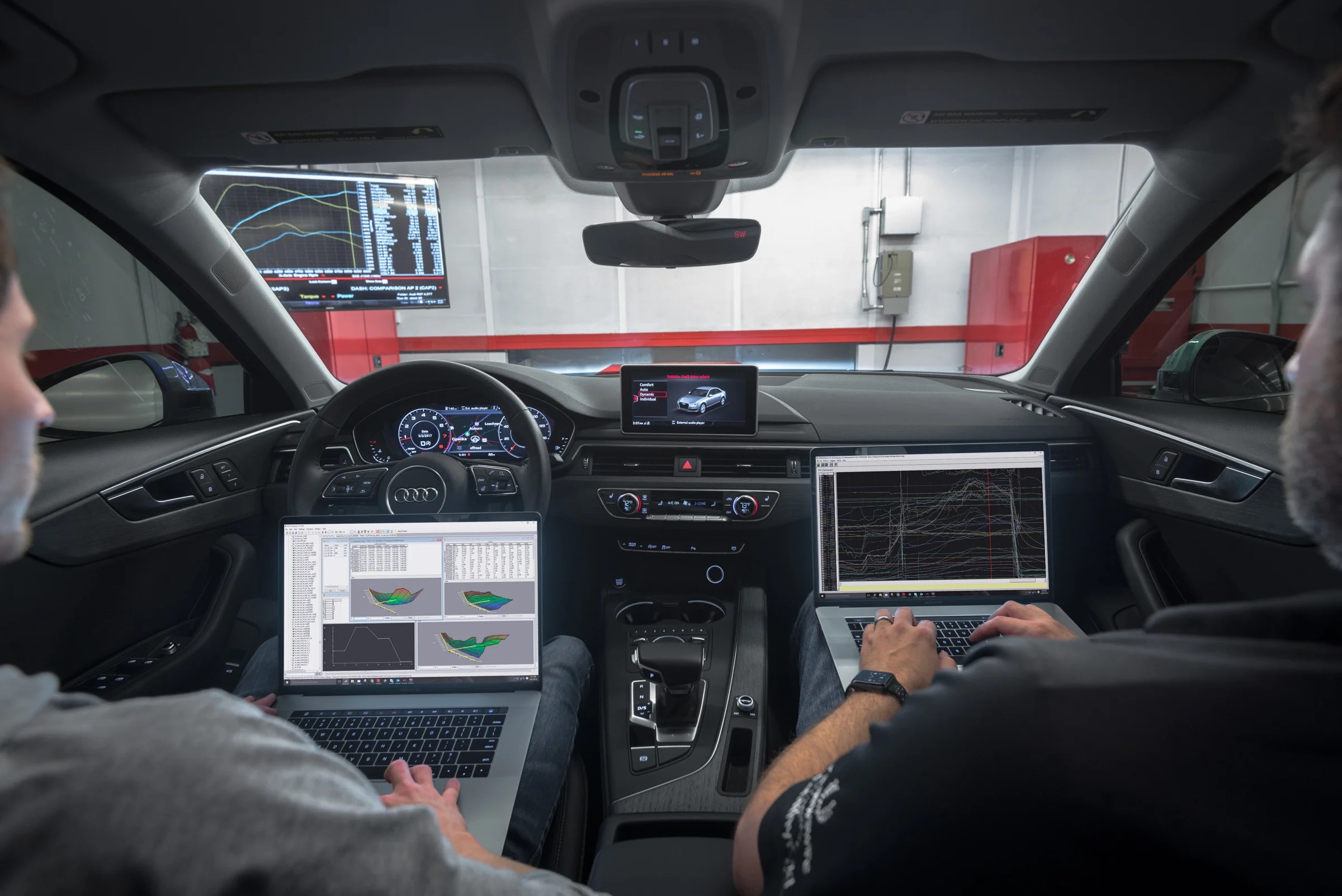 Two men with laptops sit in the front seats of a car