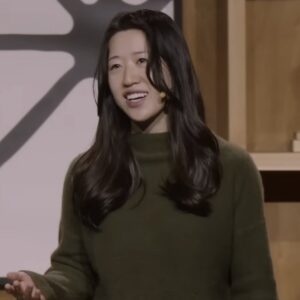 A woman with long black hair and a green sweater gestures and speaks in front of a wooden backdrop