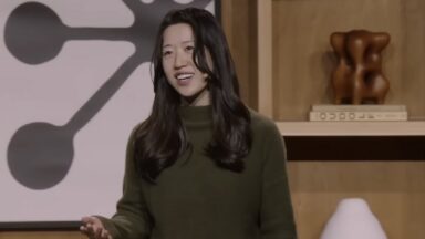 A woman with long black hair and a green sweater gestures and speaks in front of a wooden backdrop