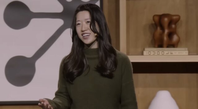 A woman with long black hair and a green sweater gestures and speaks in front of a wooden backdrop