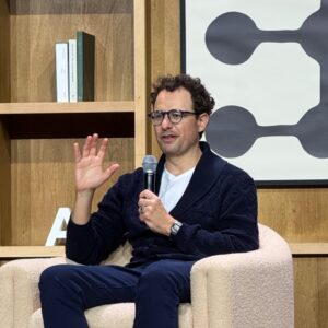 Dario Amodei sits on a chair on stage and gestures as he talks.