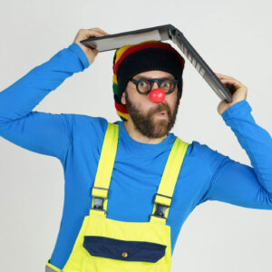 A clown in bright clothes holds a laptop above his head.