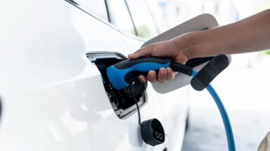 A parking lot with charging stations for electric cars. A hand holds a charging handle while charging an electric car at a home charging station.
