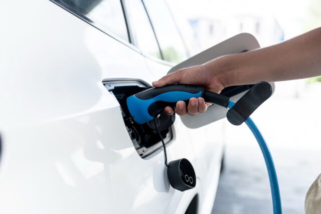 A parking lot with charging stations for electric cars. A hand holds a charging handle while charging an electric car at a home charging station.