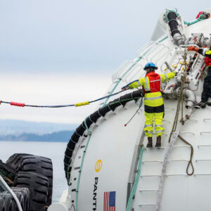 Two people stand on ladder rungs on a large, white sphere floating in the water as a third person standing on the dock or a nearby ship helps them handle a long cable. The white sphere is a prototype wave energy converter and floating AI "node" developed by the company Panthalassa.