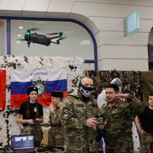 Shaman, a 19-year-old drone operator for the Russian military, flies a quadcopter drone during a demonstration event organized by members of the Berkut Military-Sports Cossack Club in a shopping centre in Voronezh, Russia on January 24, 2026. Shaman is wearing a face mask and military uniform with a camo pattern while standing next to another man in similar attire, controlling a quadcopter drone hovering in the foreground. Behind them are two girls and a boy wearing military camo-style pants and black t-shirts, along with the Russian flag.