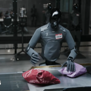 A humanoid robot with a gray body and black, reflective head stands next to a package sorting station while moving boxes and padded envelopes onto a conveyor belt.