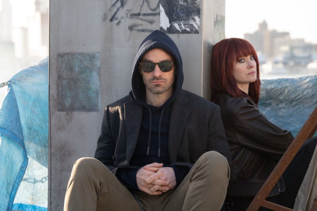 man in dark glasses and hoodie seated at the base of a column with a woman in a red wig off to the side